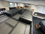 Large black storage cases are mounted on a perforated roof rack, possibly for an RV rental or adventure vehicle, in a workshop setting.