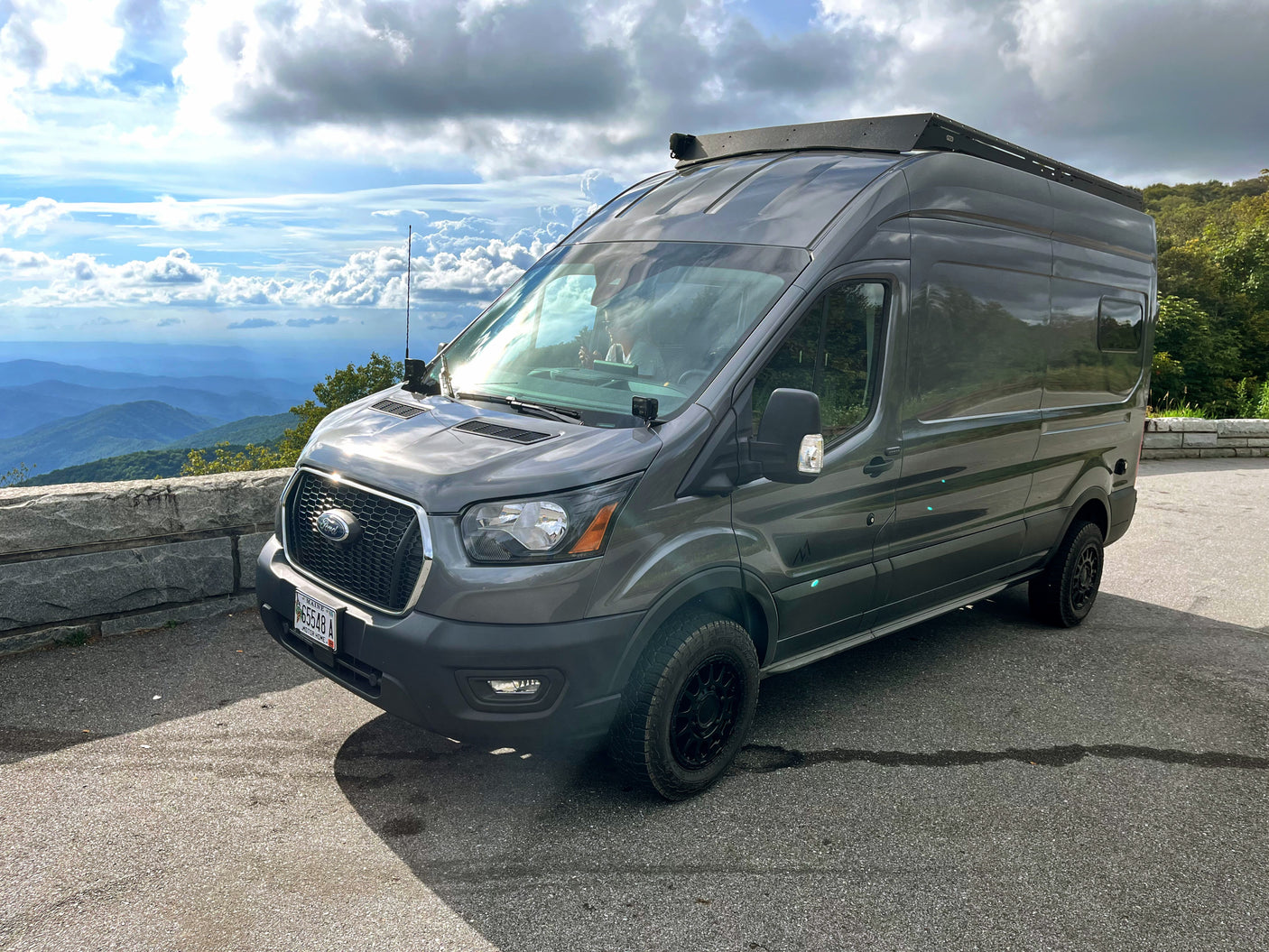 Dark gray Ford Transit van, converted to an RV rental, parked at a scenic mountain overlook under cloudy skies.