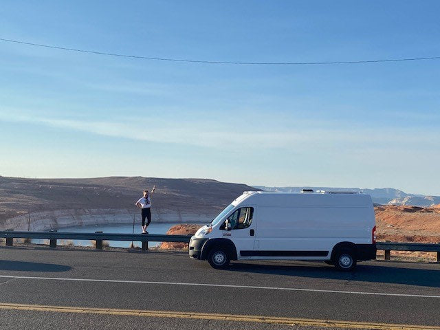 White RV rental van at a scenic lake overlook with desert cliffs. Person standing on guardrail
