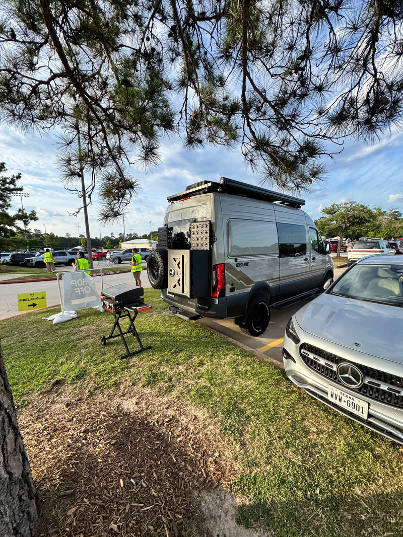 A grey Storyteller Overland 'Beast Mode' campervan next to a Mercedes-Benz car. RV rental drop-off area with a portable grill and event signs. — photo 12