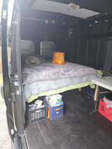 RV rental van interior showing a bed, under-bed storage crates with supplies, and a folding table setup for travel.