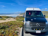 White & black RV rental Sprinter van with bear paw decals parked by a California coastal beach.