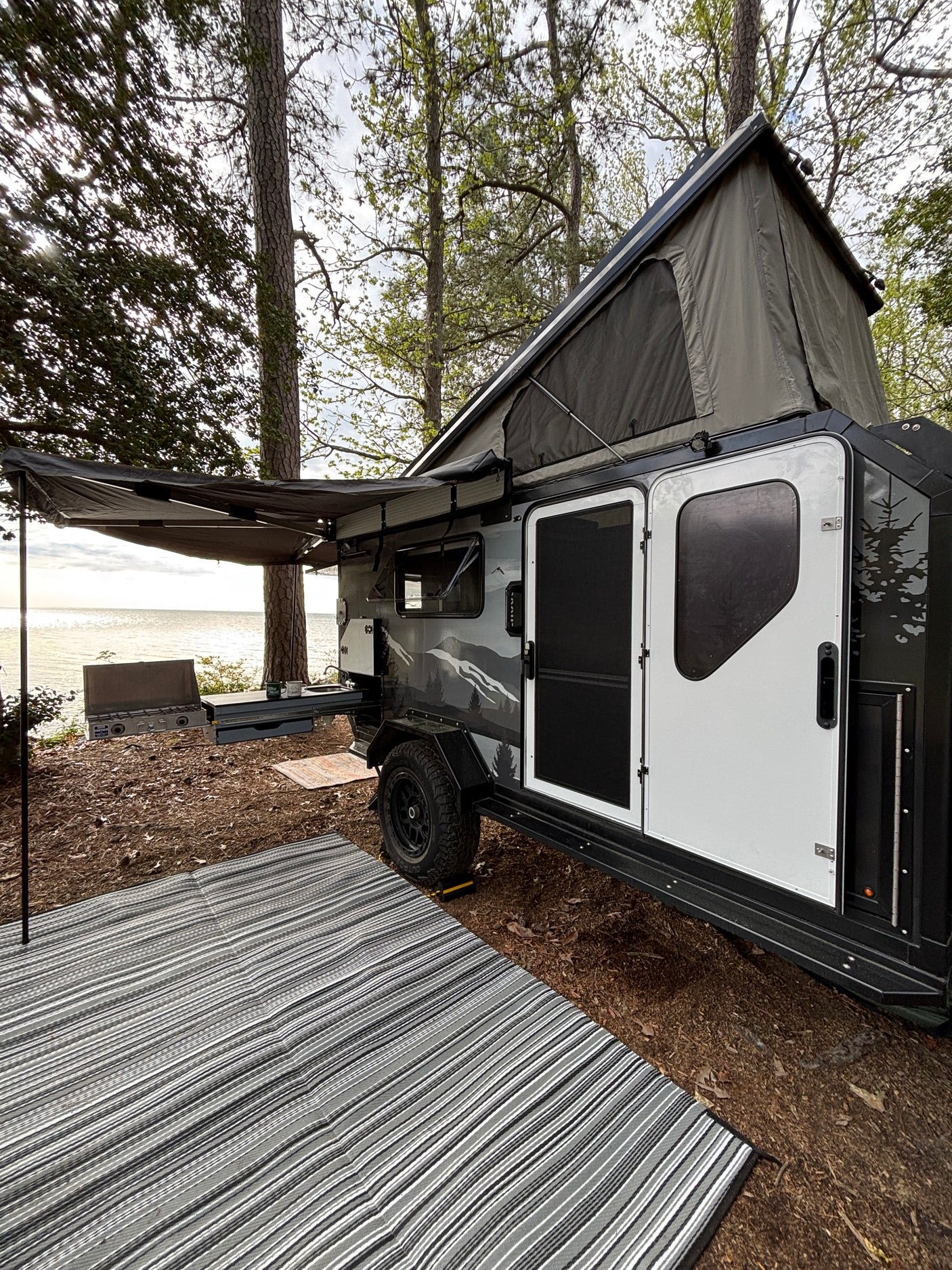 Serene lakeside camping with an RV rental. This off-road camper features a rooftop tent, awning, and outdoor kitchen by the water.