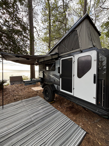 Serene lakeside camping with an RV rental. This off-road camper features a rooftop tent, awning, and outdoor kitchen by the water.