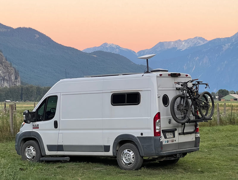 RV rental with bikes and Starlink dish in a grassy field, backed by snow-capped mountains under an orange-pink sunset sky. — photo 1