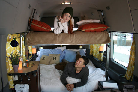 Cozy RV rental interior shows a woman on an upper bunk and a man on the lower bed inside a custom camper van.