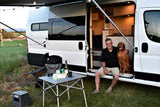 RV rental scene: A man and his dog relax at sunset by their camper van with an awning, outdoor table, and string lights.