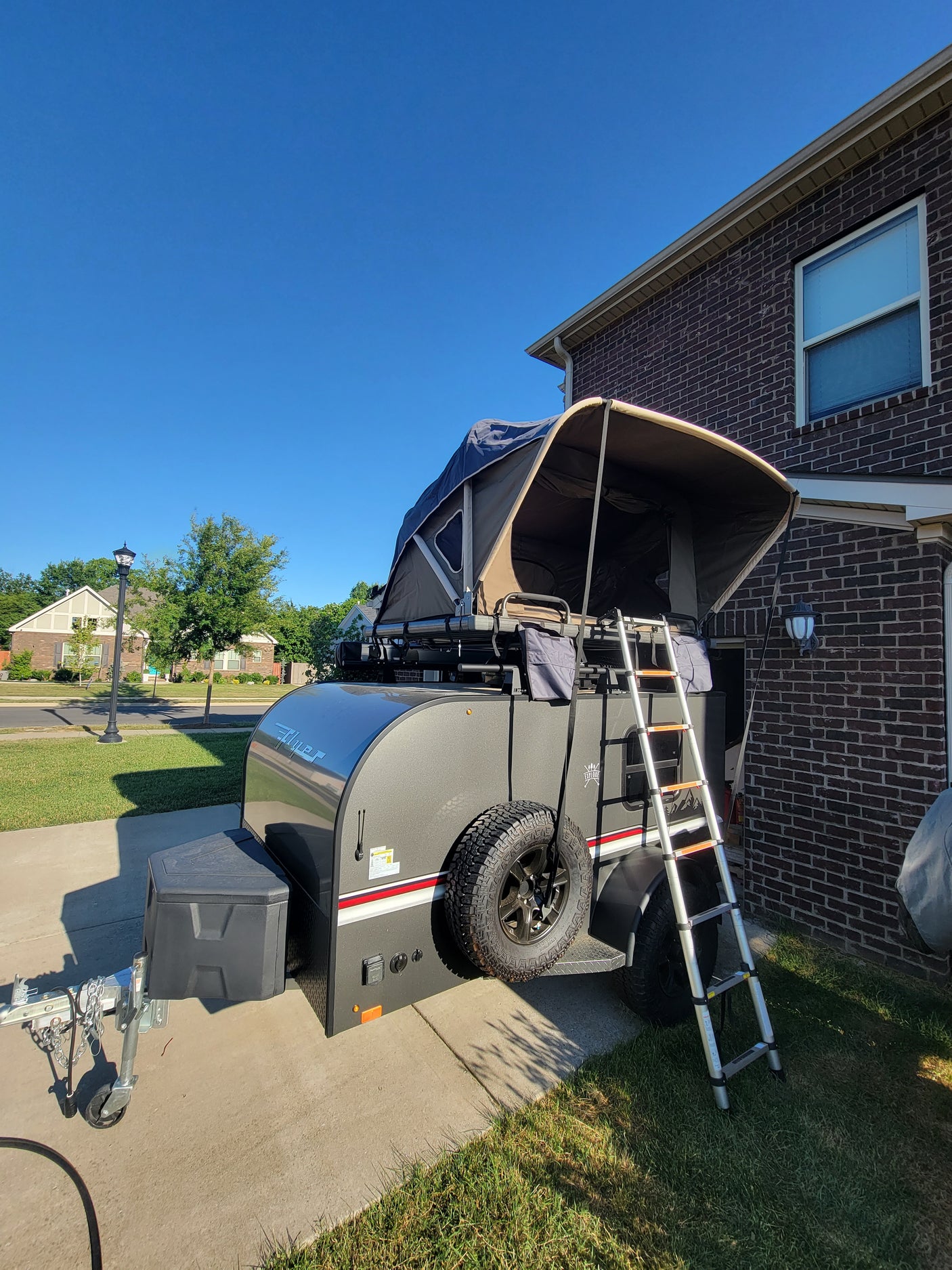 An RV rental, an off-road travel trailer with an extended awning, parked on a driveway beside a brick house.
