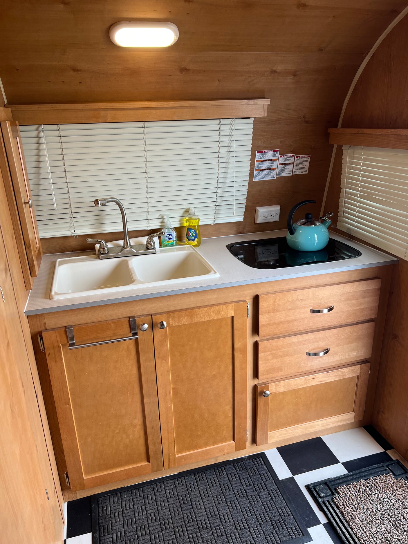 Functional RV rental kitchen with a double sink, modern stovetop, wood cabinetry, bright light, and checkered floor.