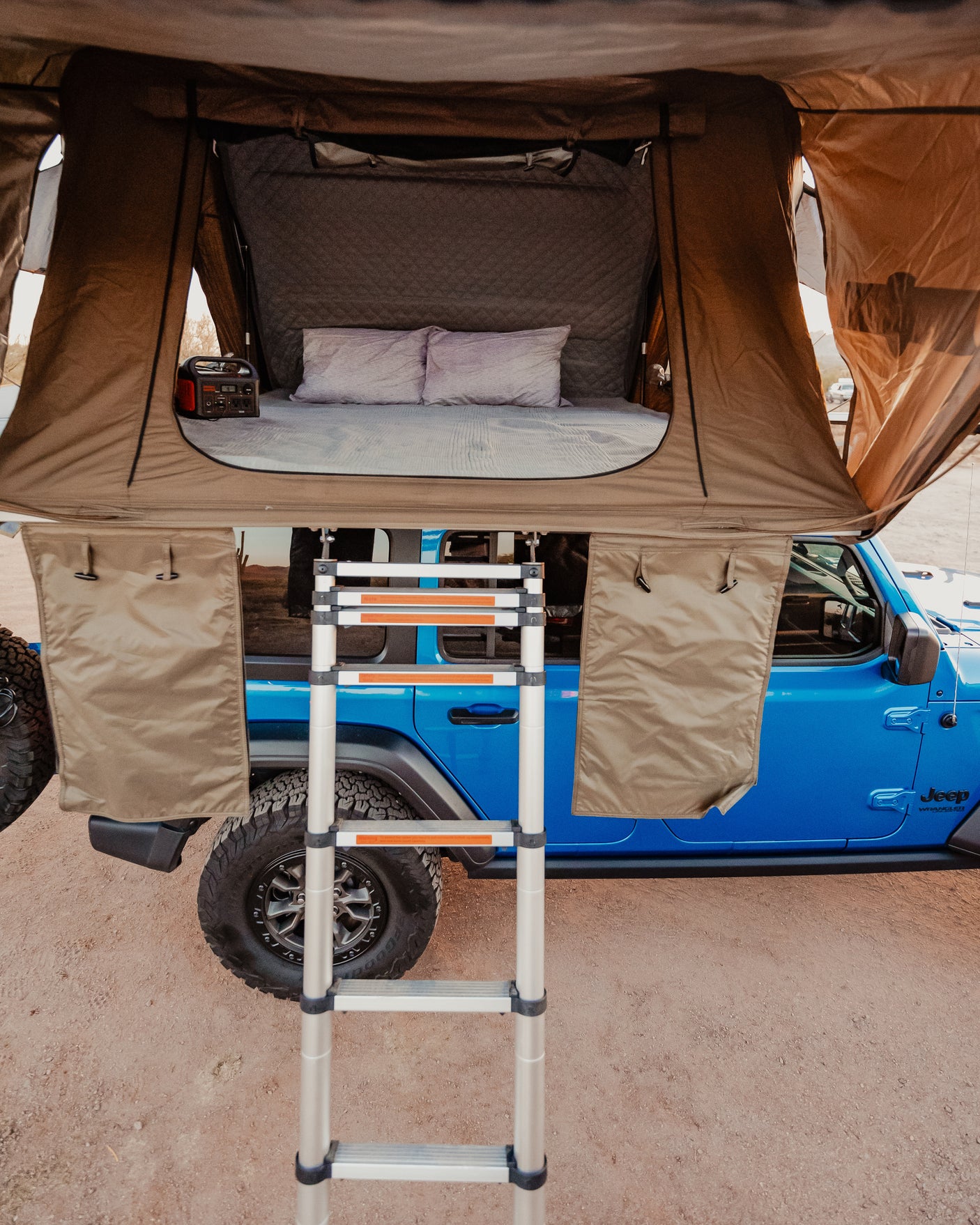 A blue Jeep with a deployed rooftop tent, featuring a bed and power station, offering an RV rental adventure.