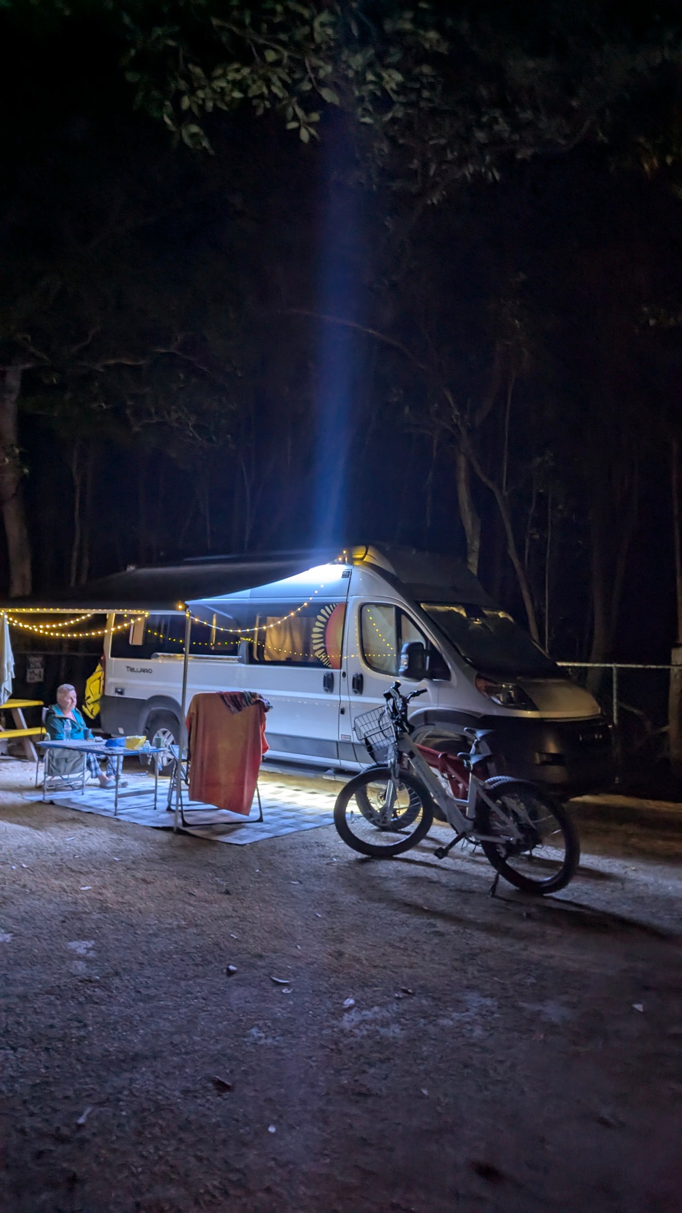 An RV rental camper van illuminates a nighttime camp. A person sits at a table with string lights, bikes parked close.