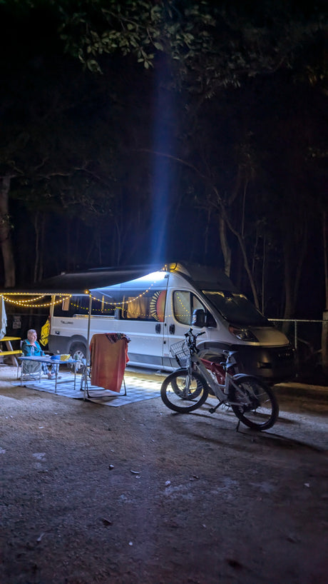 An RV rental camper van illuminates a nighttime camp. A person sits at a table with string lights, bikes parked close.