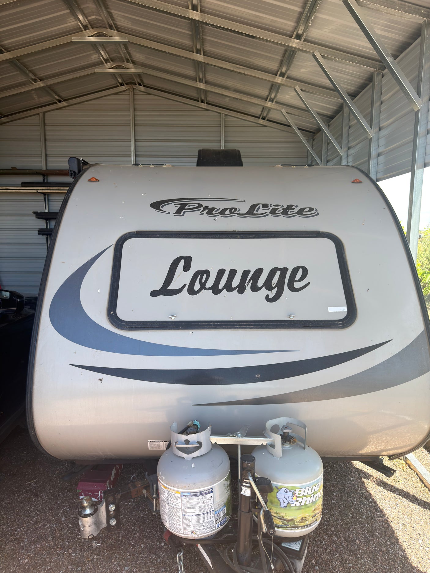Front of a ProLite 'Lounge' travel trailer with propane tanks and hitch, safely stored under a carport. Perfect for rv rental.