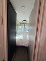 View down a narrow RV rental interior showing a cozy white bed at the end, flanked by a refrigerator and wood-grain cabinets.