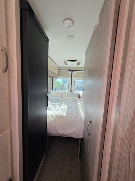 View down a narrow RV rental interior showing a cozy white bed at the end, flanked by a refrigerator and wood-grain cabinets.