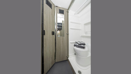 Compact RV rental bathroom: wood-grain walls, toilet, shower, mirror on door. Efficient space for travelers.