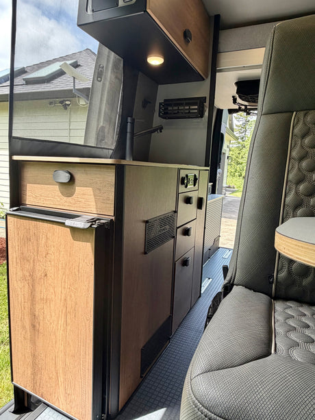 Modern rv rental interior with kitchen, fridge, cabinets, and seating. View outside through screen shows house and sky.