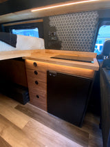 Modern RV rental interior featuring a cozy bed, wood kitchenette with fridge, cooktop, and stylish hexagon tile backsplash.