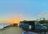 Blue RV rental camper van on sandy bluff, sunset over ocean & waves. Sun low on horizon.