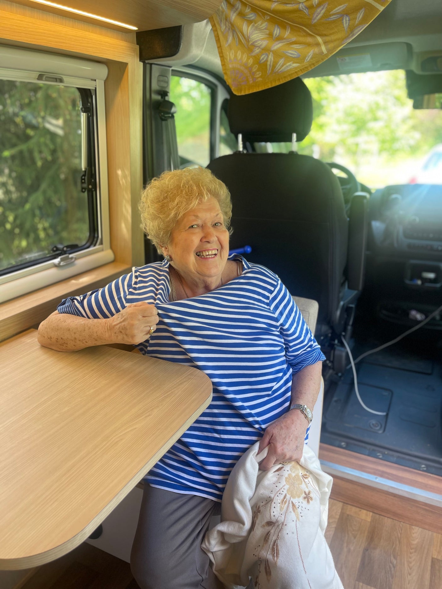 A cheerful senior woman smiles inside a modern RV rental, sitting by the wooden table and window.