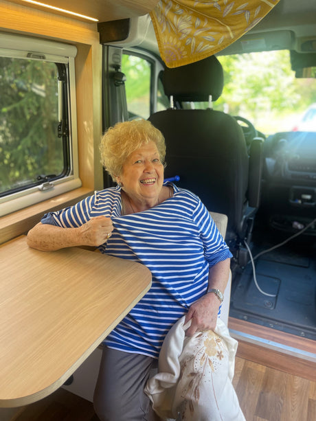 A cheerful senior woman smiles inside a modern RV rental, sitting by the wooden table and window.