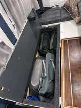 RV rental interior featuring open under-bed storage with camping gear, bags, and folded items. Cushions are visible.