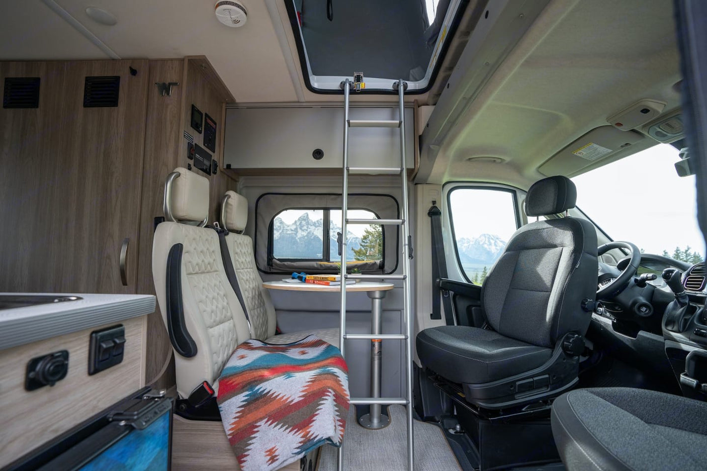 Inside a comfortable RV rental: swiveling seats, dining table, kitchen, and a ladder to a bunk, all with scenic mountain views.