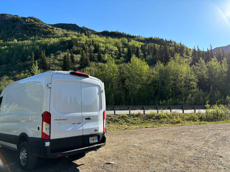 Scenic Alaskan view: a white Ford Transit RV rental van parked on a dirt pull-off by a lush green mountain road.