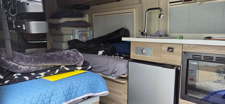 RV rental interior: bed, stacked pillows/bedding, and kitchenette with sink, eco-HOT control, fridge, and microwave.