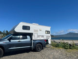 A blue truck with a white camper, ideal for an RV rental, is parked on a gravel lot by a bay with distant mountains.