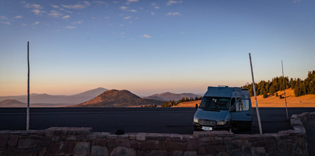 Blue camper van, ideal as an rv rental, parked at a scenic mountain overlook with a stone wall and golden light.