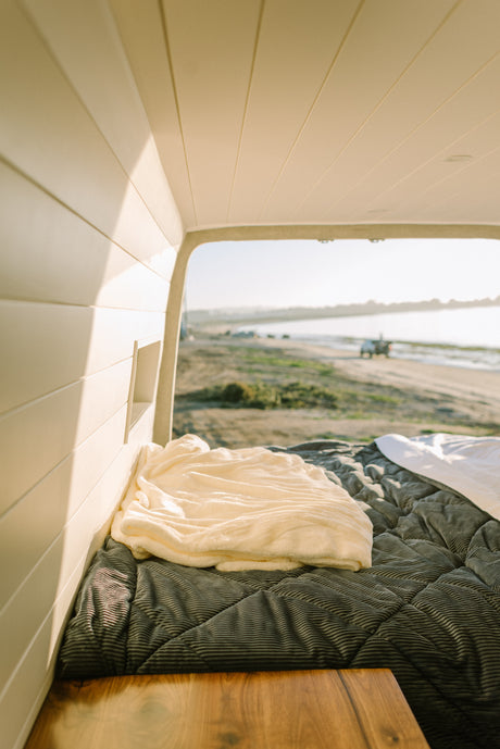 Cozy RV rental interior: a soft bed overlooks a serene beach scene, perfect for relaxing travel.