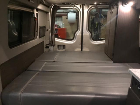 Inside an RV rental: a modern camper van with flexible grey bed cushions, integrated storage, and wide windows.