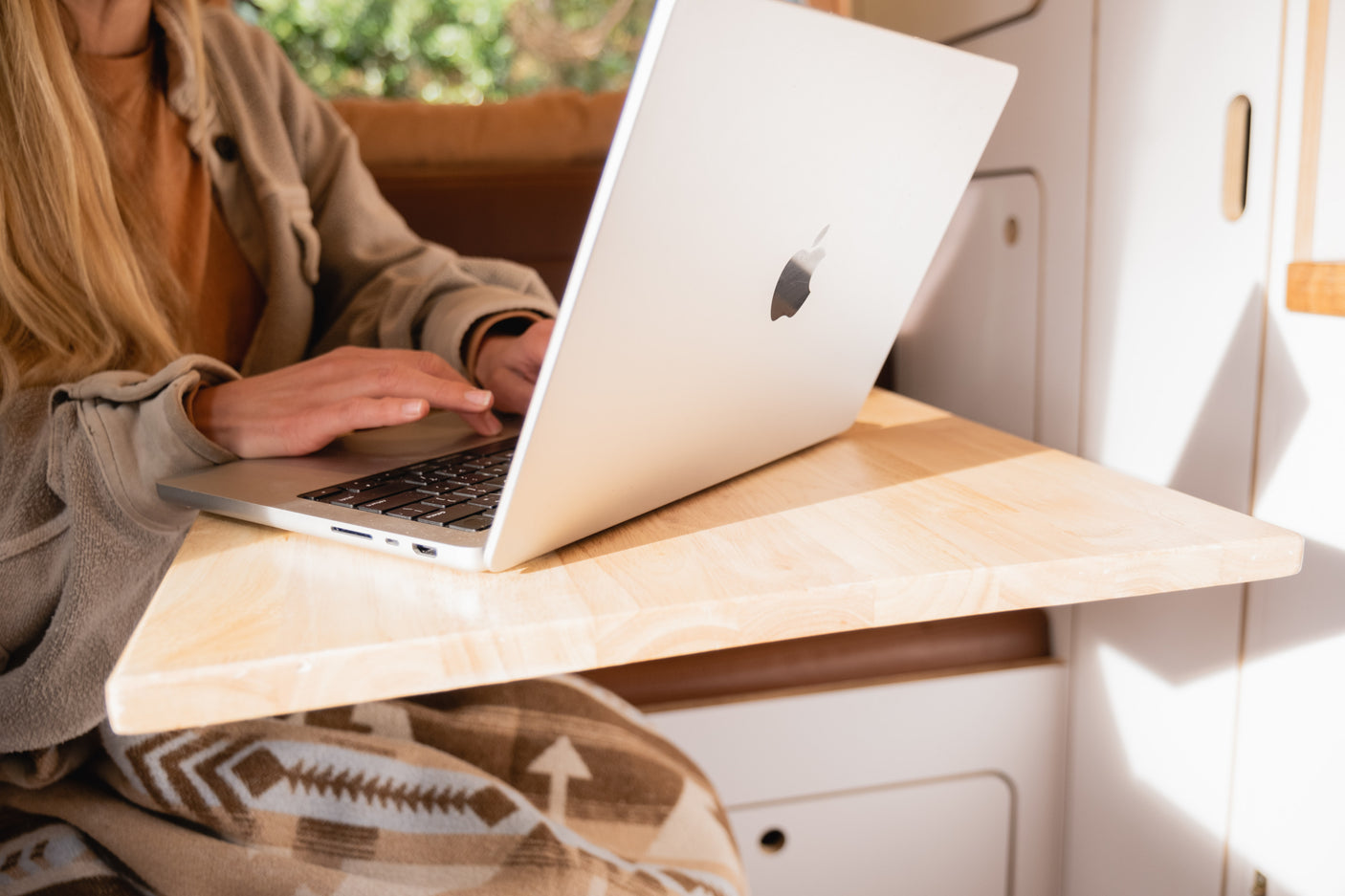Working remotely in an RV rental. A person uses a MacBook on a wooden table. A cozy patterned blanket covers their lap.