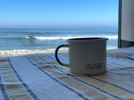 Morning coffee by the beach. This RV rental offers stunning ocean views, with a white 'TEXINO' mug on a striped blanket.