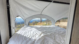 Inside an RV rental, a comfy bed faces panoramic mountain views from a pop-up tent extension. A campsite is visible outside.