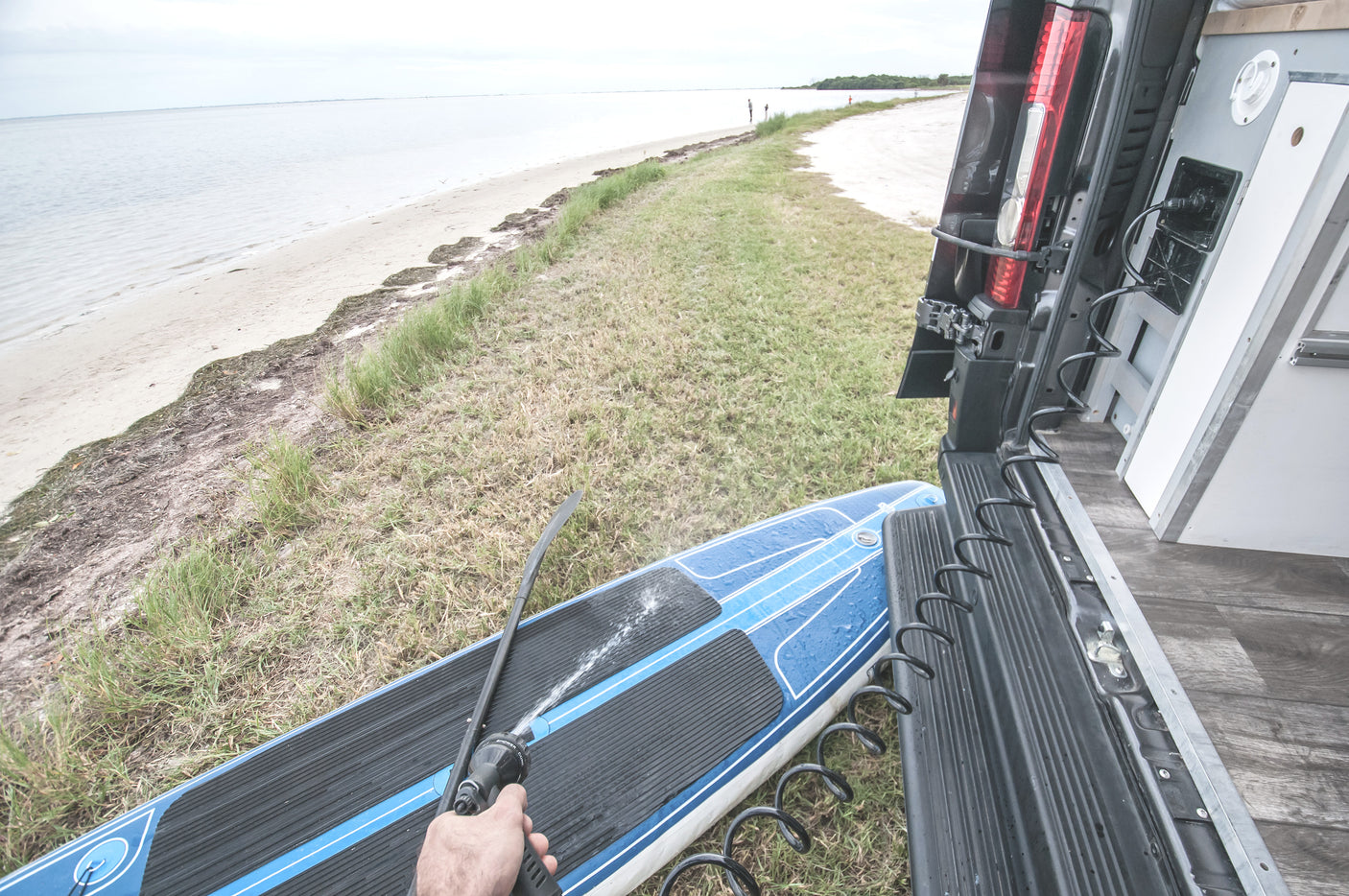 A person sprays a blue paddleboard with water from a hose connected to a parked RV rental, seaside. Ideal for post-water cleanup.