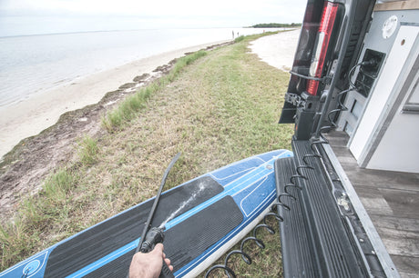 A person sprays a blue paddleboard with water from a hose connected to a parked RV rental, seaside. Ideal for post-water cleanup.