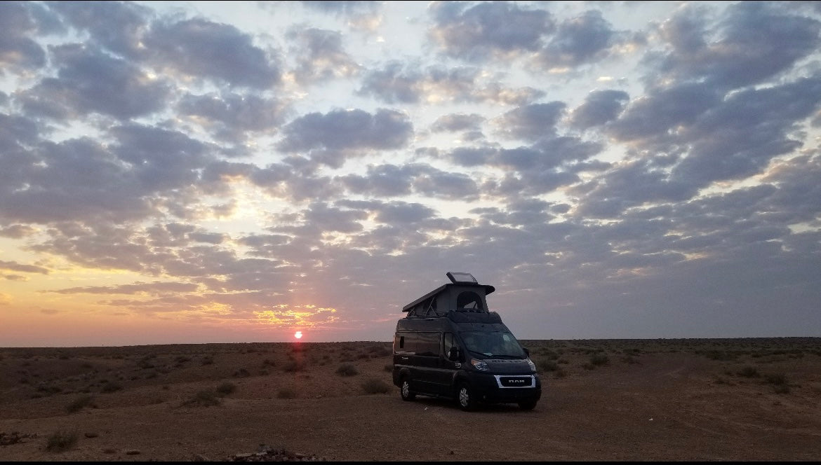 Black rv rental parked in a desert at sunset, pop-top roof extended, under a cloudy sky.