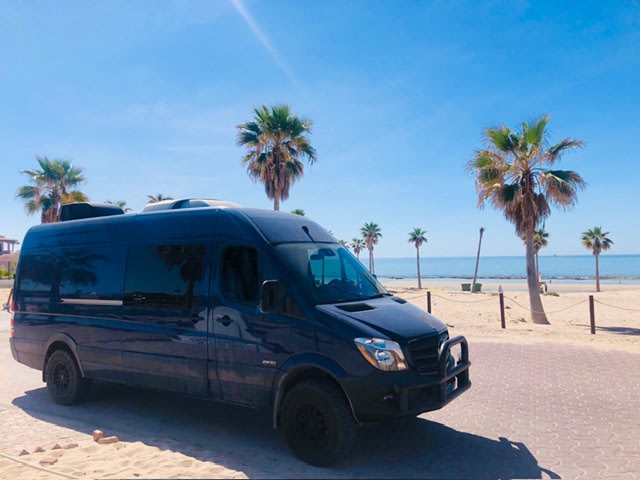 Explore coastal beauty with this dark blue RV rental. A camper van parked on a sunny beach, surrounded by palm trees & ocean. — photo 3