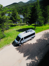 Overhead view: A light grey, solar-equipped RV rental parked on a road, overlooking a serene lake and dense pine forest.