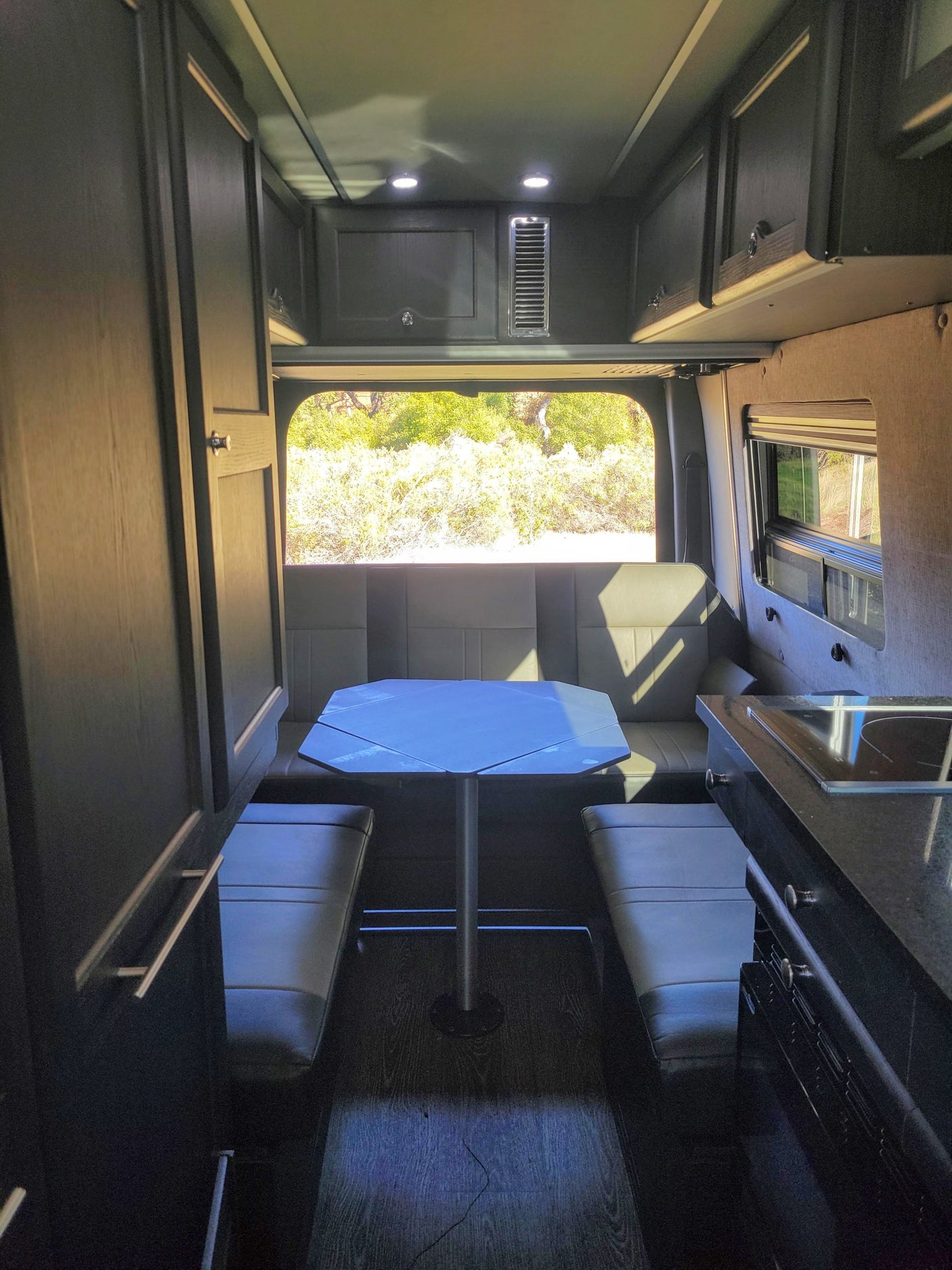 An RV rental interior showcasing a dining area with a table, bench seating, and a large rear window. Kitchen on the right.
