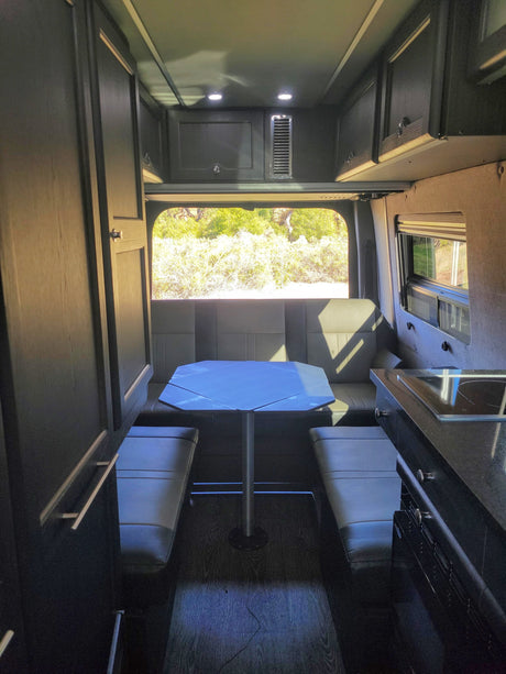 An RV rental interior showcasing a dining area with a table, bench seating, and a large rear window. Kitchen on the right.