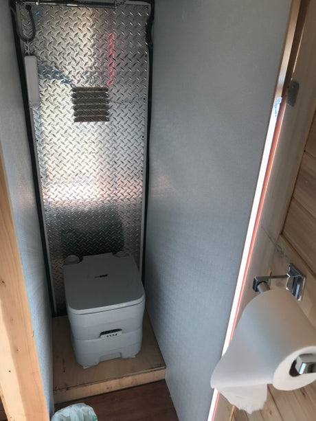 Compact RV rental bathroom featuring a portable toilet, diamond plate wall, and toilet paper dispenser. Ideal for travel.
