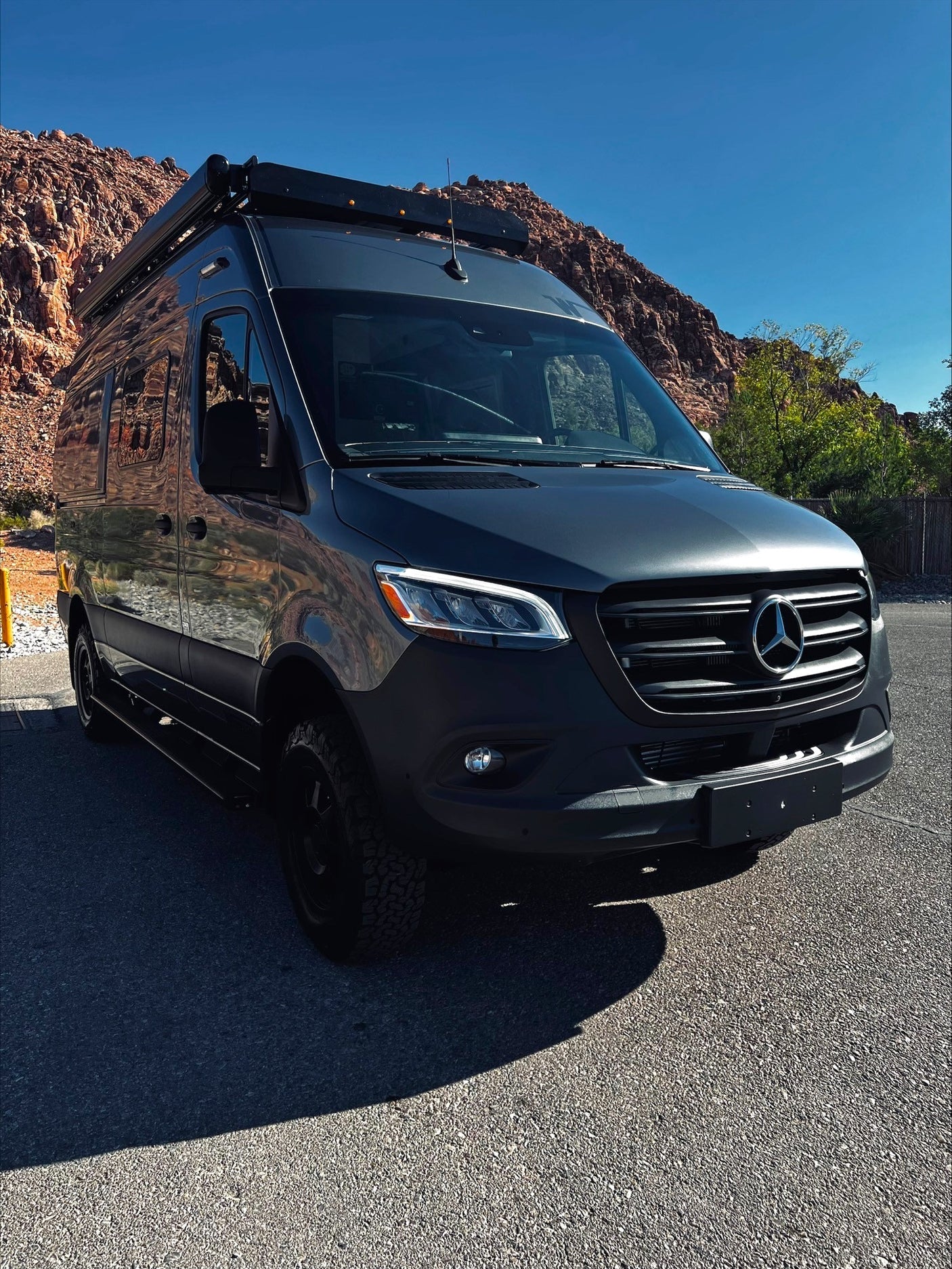 A dark gray Mercedes Sprinter RV rental, parked on asphalt with red rocky mountains and a bright blue sky in the background.