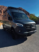 A dark gray Mercedes Sprinter RV rental, parked on asphalt with red rocky mountains and a bright blue sky in the background.