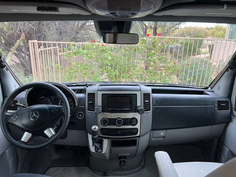 Front cabin of a clean Mercedes-Benz Sprinter RV rental, showing the dashboard, steering wheel, and a view outside.