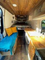 Inside this RV rental, a cozy van interior reveals a wood ceiling, rear bed, side bench, and live-edge kitchen.