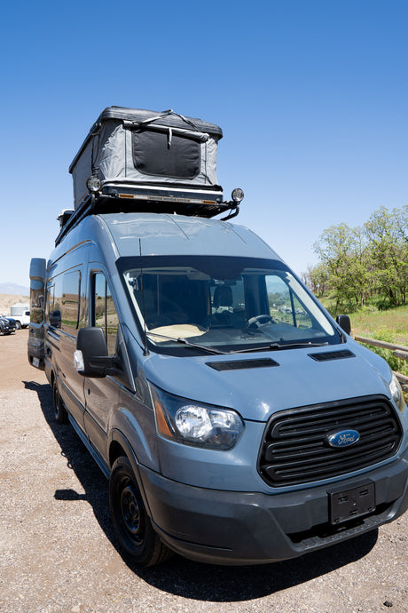 A gray Ford Transit campervan with a rooftop tent, parked on a gravel road under a clear sky. Ideal for RV rental adventures.