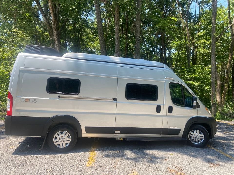 Experience nature with this Winnebago Solis RV rental, a sleek camper van parked among green trees on an asphalt lot.